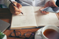 Hands holding a phone and pen, with open diary, glasses and cup of tea on the table